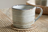 The nkuku Vizo Mug - Grey - Large features subtle blue-gray and tan tones, shown on a woven coaster atop ribbed wood, with a matching tray and another mug softly blurred in the background.