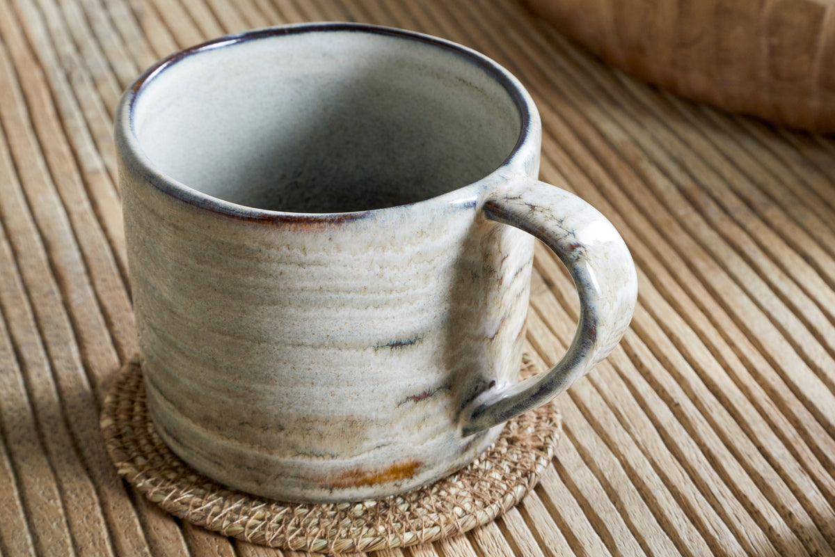 The Vizo Mug - Grey - Large by nkuku, featuring subtle brown and gray tones, rests on a round woven coaster atop a grooved wooden table.