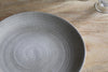 A close-up of the nkuku Valinha Earthenware Dinner Plate in grey, featuring a reactive glaze, sits empty on a light wooden table with the edge of a clear glass visible in the upper right corner.
