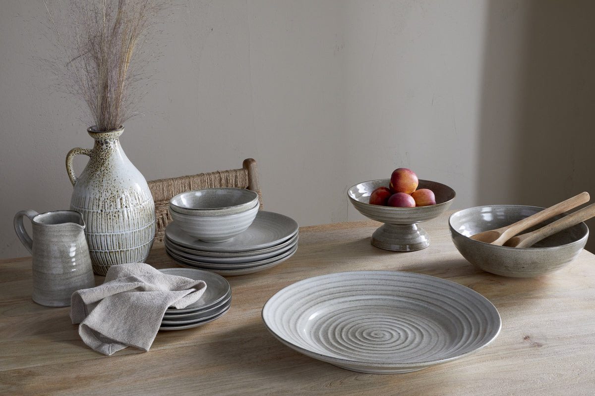 A rustic wooden table is set with the nkuku Valinha Earthenware Dinner Plate in grey, ceramic bowls, a jug, vase of dried grasses, a bowl of peaches, wooden utensils, and a folded linen napkin for a charming tableware collection against a neutral wall.