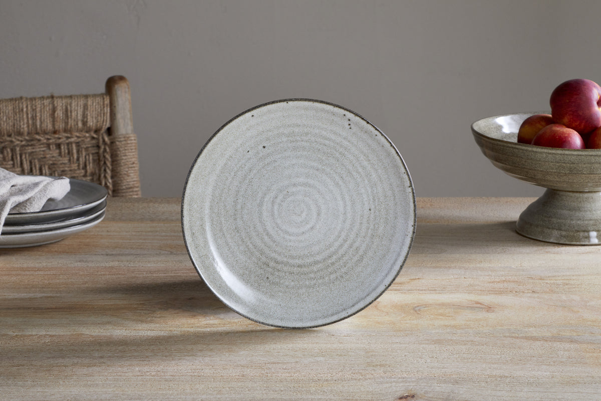 The Valinha Earthenware Dinnerware Set - Grey by nkuku features a light gray ceramic plate with circular patterns and reactive glaze, standing on a wooden table beside stacked plates, a woven chair, and a fruit bowl with red apples.