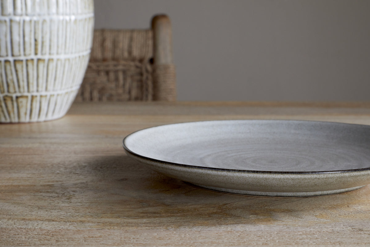 The Valinha Earthenware Side Plate - Grey by nkuku rests on a wooden table, accompanied by part of a textured vase and a woven chair in the background, creating a calm and minimalistic setting.