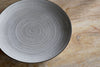 The Valinha Earthenware Side Plate in grey by nkuku features a textured spiral design and round shape, displayed on a light wooden surface to highlight its distinctive tableware charm.