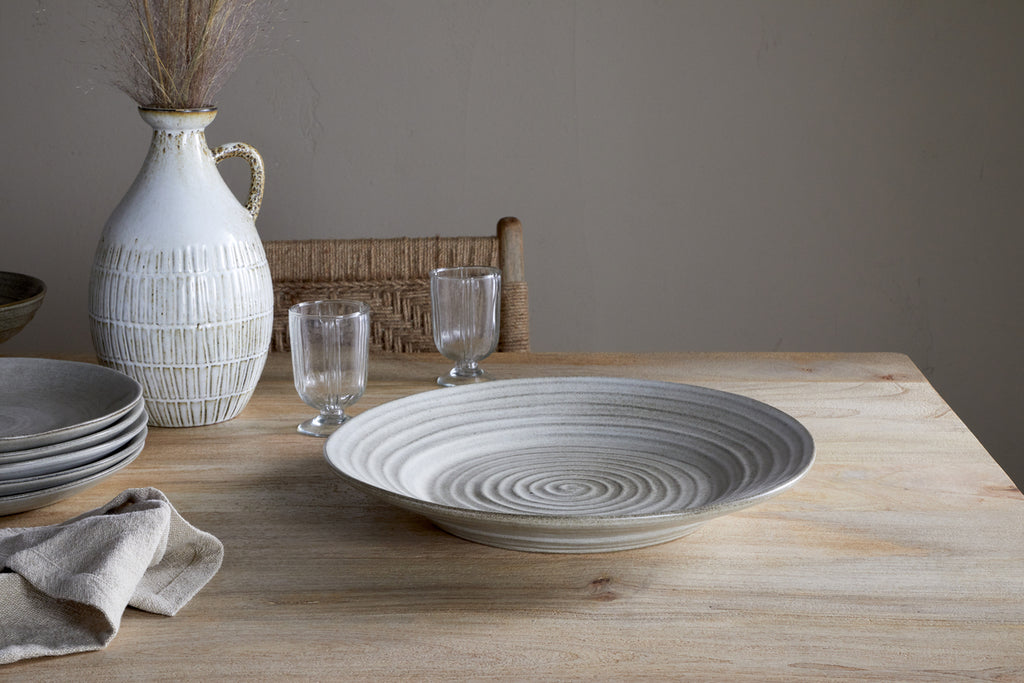 The Valinha Earthenware Round Platter in grey by nkuku rests on a wooden table, paired with two clear glasses, a white vase with dried grass, stacked plates, a folded beige napkin, and a woven chair in the background.