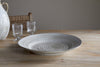 The Valinha Earthenware Round Platter in Grey by nkuku rests on a wooden table alongside two clear glass goblets and a white patterned jug, with a woven chair partially visible in the background.