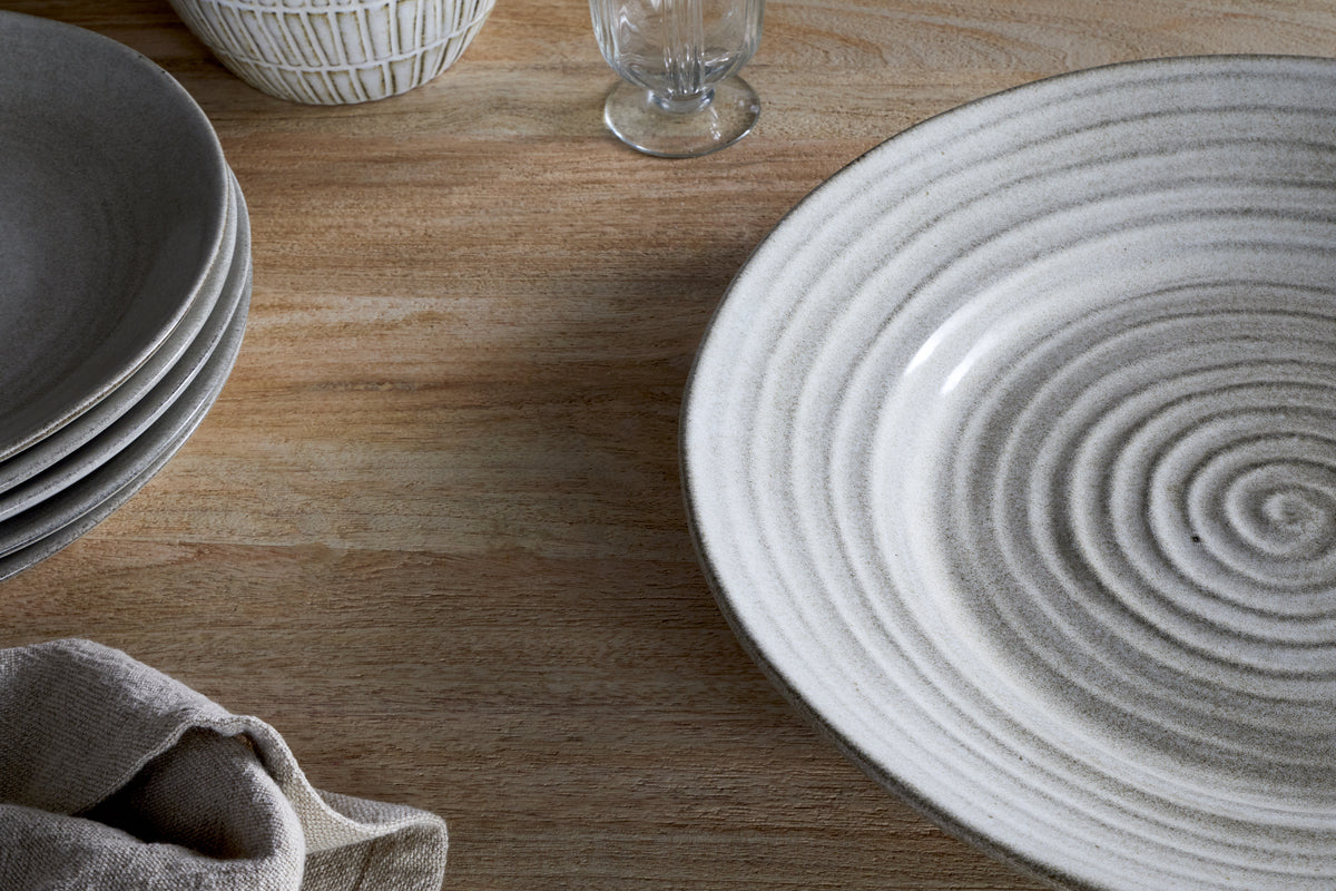 A close-up of a wooden table set with the nkuku Valinha Earthenware Round Platter in grey, stacked serveware, a linen napkin, a decorative bowl, and a glass—all in neutral hues with an eye-catching reactive glaze.
