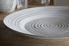 The Valinha Earthenware Round Platter in grey by nkuku, featuring concentric ridges, rests on a wooden table with glassware and textured ceramic serveware in the background.