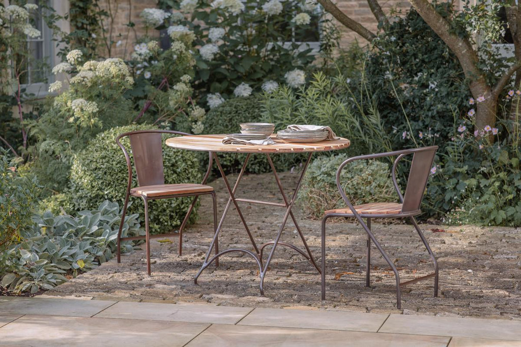 The nkuku Vivika Outdoor Dining Table - Natural sits on a stone patio, paired with two matching metal chairs with wooden seats. Stacked plates and napkins rest on the table, while lush greenery and white flowers fill the garden backdrop.