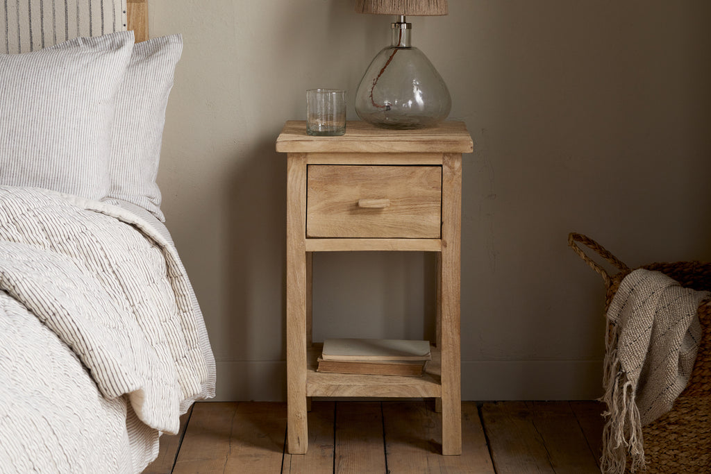The Vivan Grooved Bedside Table - Natural by nkuku holds a glass lamp, drinking glass, and two books on its lower shelf beside a bed with neutral bedding and a woven basket on the wooden floor.