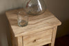 A clear glass tumbler and a large round glass vase rest on the nkuku Vivan Grooved Bedside Table - Natural, which features a drawer and sits against a neutral-colored wall.