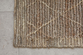 A close-up of the nkuku Vandi Jute Doormat - Natural, featuring thick, textured braids in beige and brown tones with a geometric diamond design, displayed on a light gray stone tile floor.