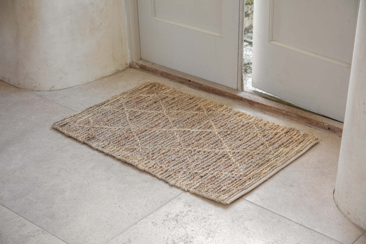 The nkuku Vandi Jute Doormat - Natural, a rectangular beige mat with a woven diamond design, sits on a light stone floor by a white door with glass panels as sunlight brightens the textured surface in the entryway.