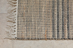 A close-up of the nkuku Varli Jute & Cotton Doormat - Natural shows black, beige, and tan checkered stripes with an off-white fringe on the left edge, resting on a light textured stone floor. The weaving looks coarse and handmade.