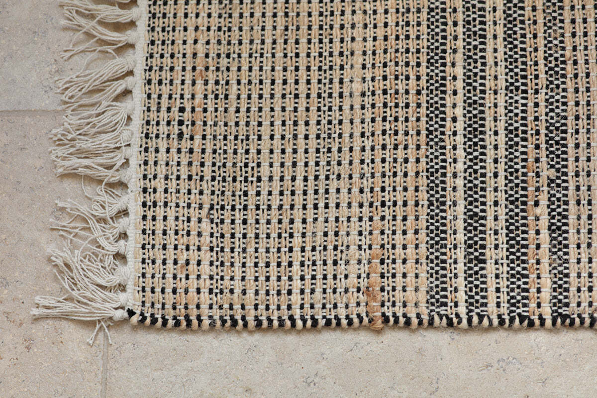 A close-up of the nkuku Varli Jute & Cotton Doormat - Natural shows black, beige, and tan checkered stripes with an off-white fringe on the left edge, resting on a light textured stone floor. The weaving looks coarse and handmade.
