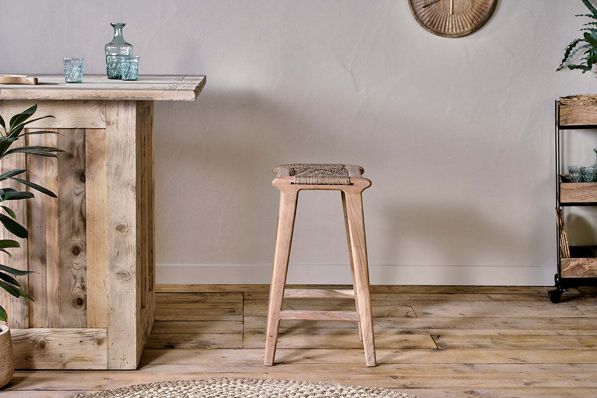 The Vinay Woven Counter Stool - Natural by nkuku, with a jute seat and acacia wood frame, is placed at a rustic wooden bar in a room with plank flooring and a textured wall finish.