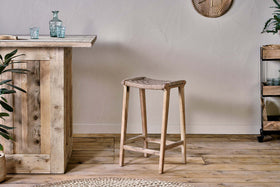 A rustic interior showcases the nkuku Vinay Woven Counter Stool – Natural at a wooden bar counter, complemented by a small metal plant shelf on the right and a round woven rug partially seen on the wooden floor.