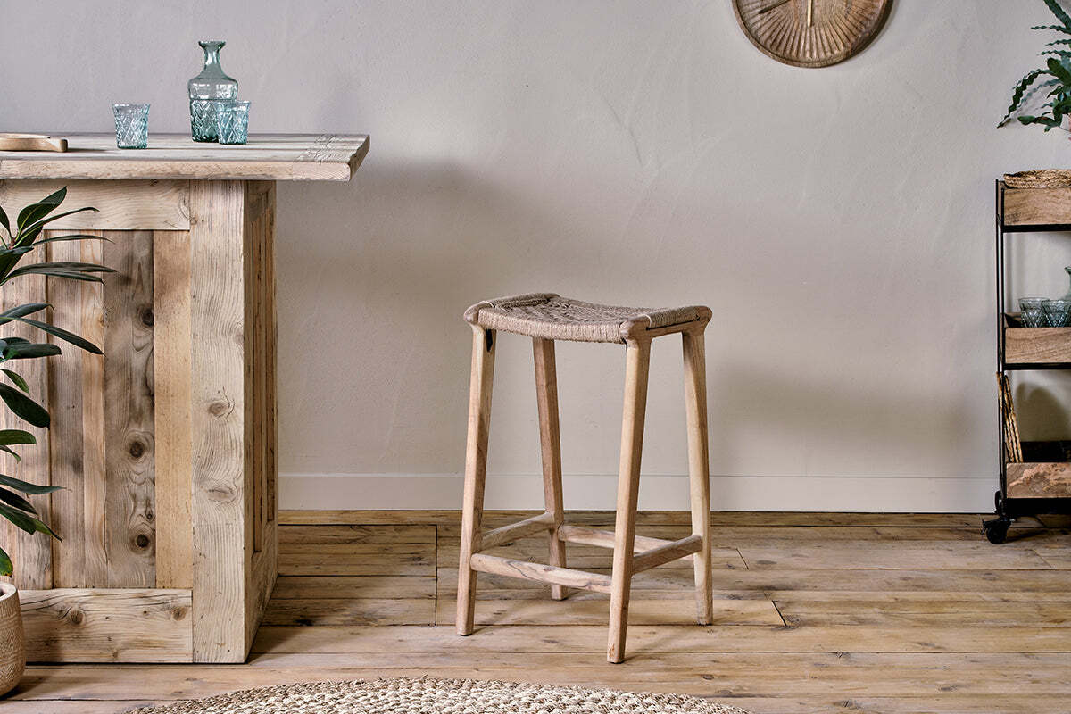 A rustic interior showcases the nkuku Vinay Woven Counter Stool – Natural at a wooden bar counter, complemented by a small metal plant shelf on the right and a round woven rug partially seen on the wooden floor.