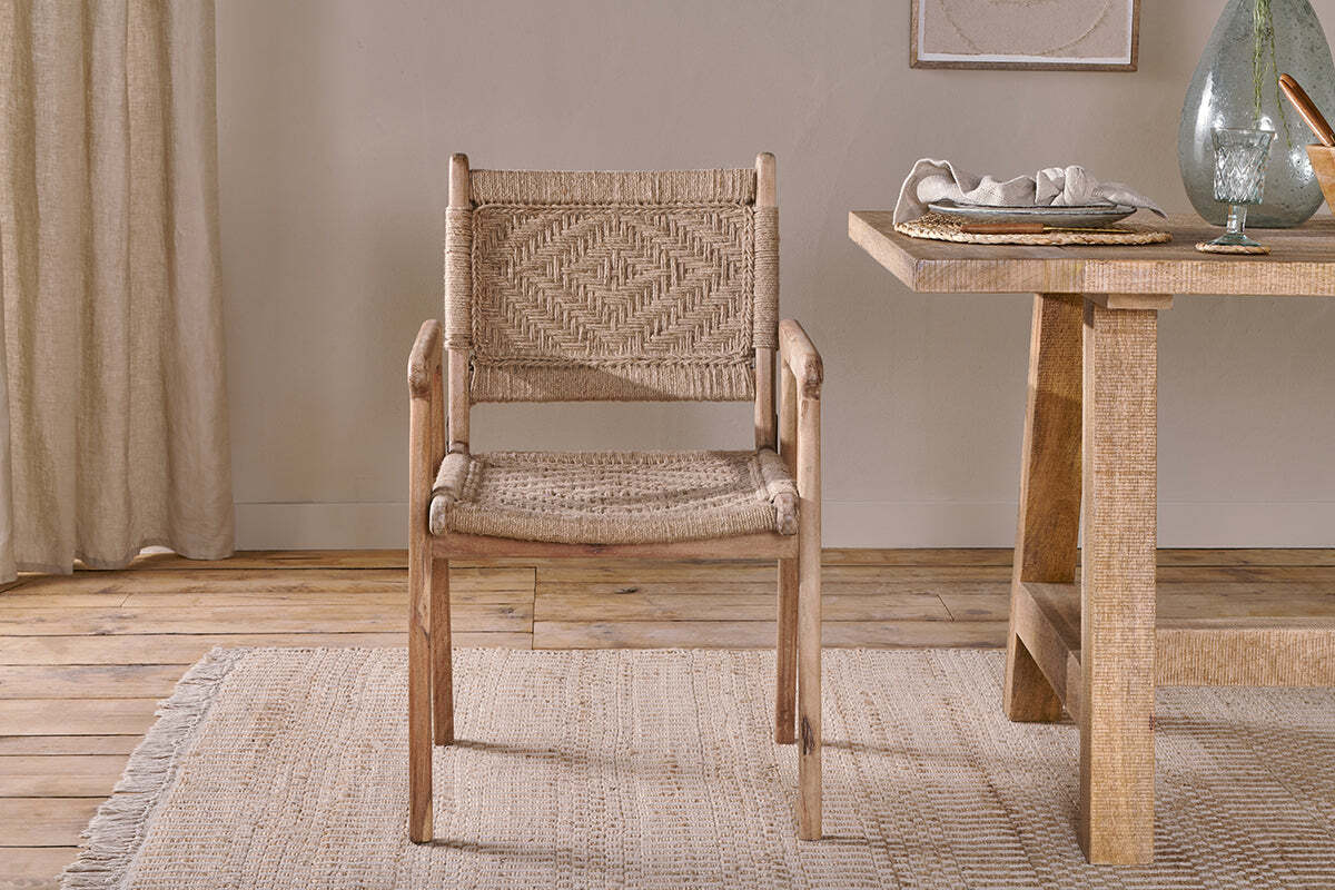 The nkuku Vinay Woven Dining Chair - Natural sits on a textured beige rug beside a wooden dining table set with dishes and glassware, surrounded by neutral decor, light wood floors, and jute paneling in the background.