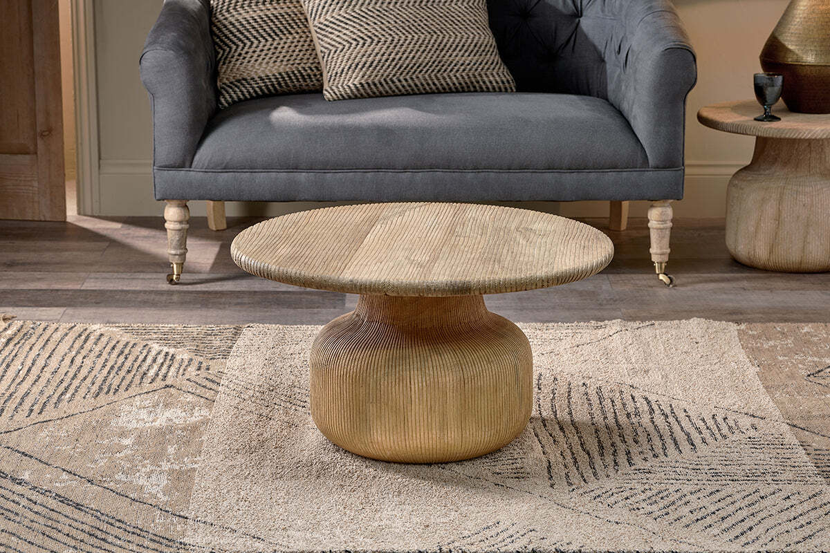 A gold, round nkuku Vivan Grooved Wood Coffee Table with a textured base and smooth top sits on a patterned beige rug, paired with a gray upholstered sofa and patterned pillows for a cozy, modern living space in neutral tones.
