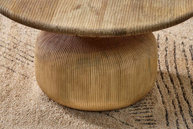 A close-up of the Vivan Grooved Wood Coffee Table by nkuku shows its vertically grooved, rounded base in sustainable mango wood supporting a thick top. The modern table stands on a beige rug with an abstract linear design.