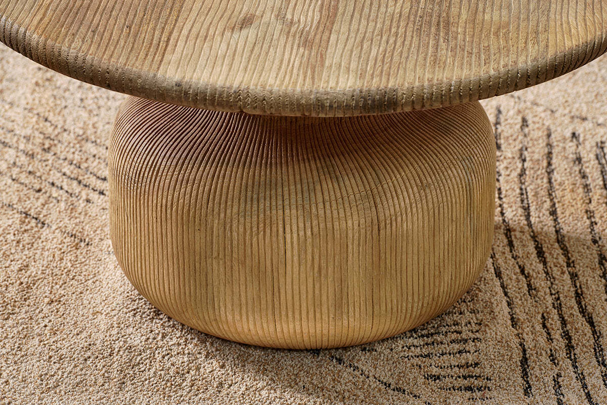A close-up of the Vivan Grooved Wood Coffee Table by nkuku shows its vertically grooved, rounded base in sustainable mango wood supporting a thick top. The modern table stands on a beige rug with an abstract linear design.