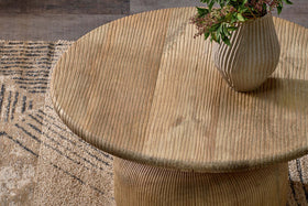 The nkuku Vivan Grooved Wood Coffee Table, crafted from sustainable mango wood, stands on a patterned beige and brown rug, topped with a ribbed ceramic vase and greenery for an earthy touch.