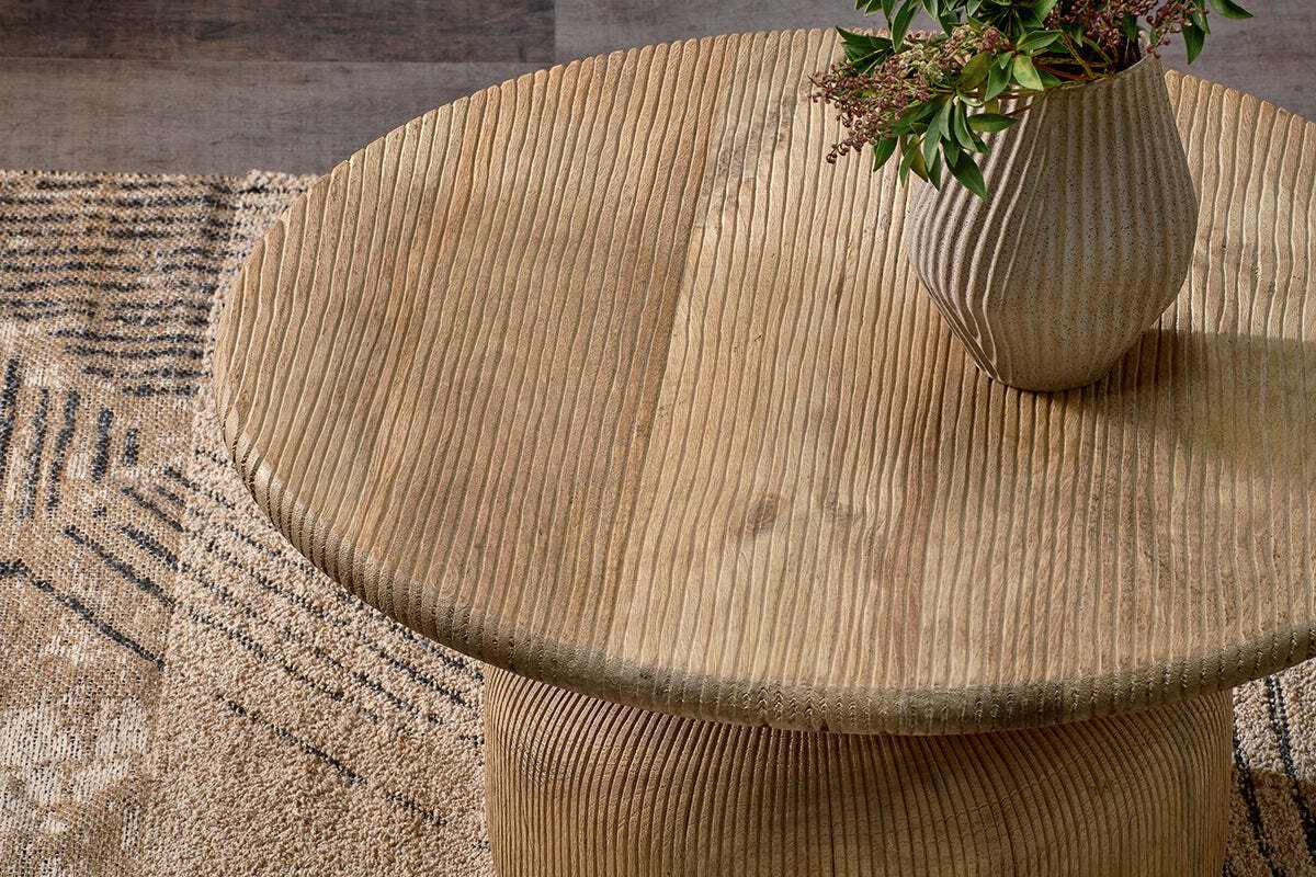 The nkuku Vivan Grooved Wood Coffee Table, crafted from sustainable mango wood, stands on a patterned beige and brown rug, topped with a ribbed ceramic vase and greenery for an earthy touch.