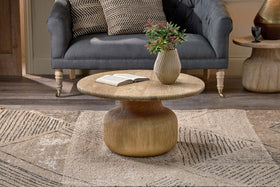 The nkuku Vivan Grooved Wood Coffee Table, made from sustainable mango wood, displays an open book and a vase with greenery. Behind it, a dark grey tufted sofa rests on a geometric beige rug over wooden flooring.