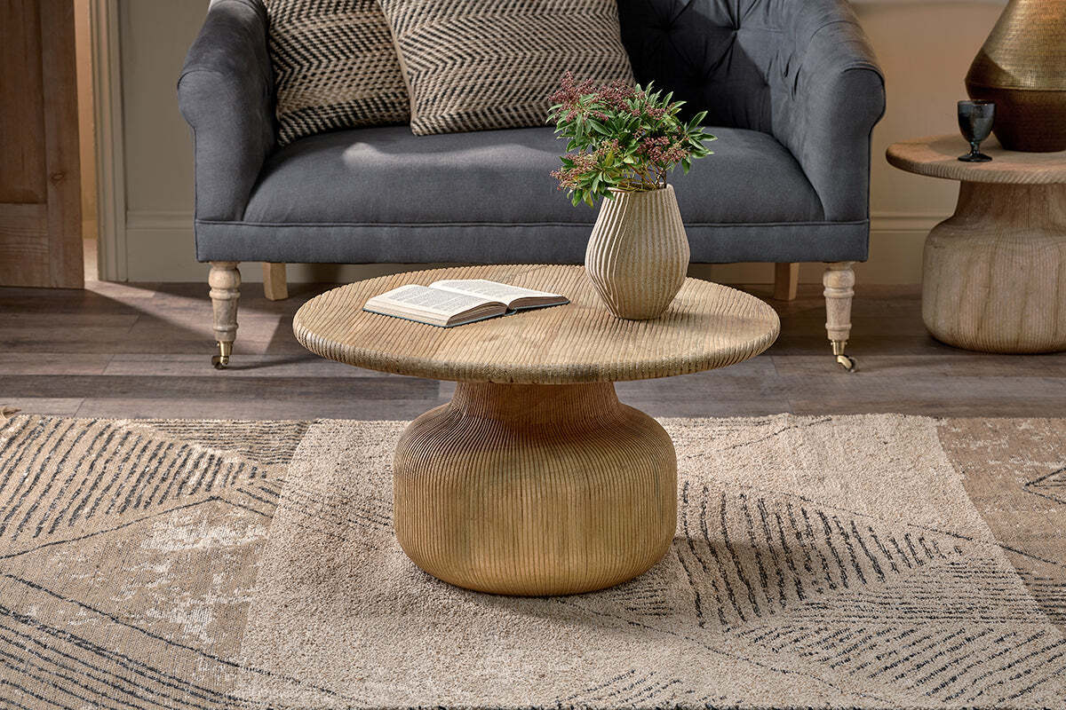 The nkuku Vivan Grooved Wood Coffee Table, made from sustainable mango wood, displays an open book and a vase with greenery. Behind it, a dark grey tufted sofa rests on a geometric beige rug over wooden flooring.