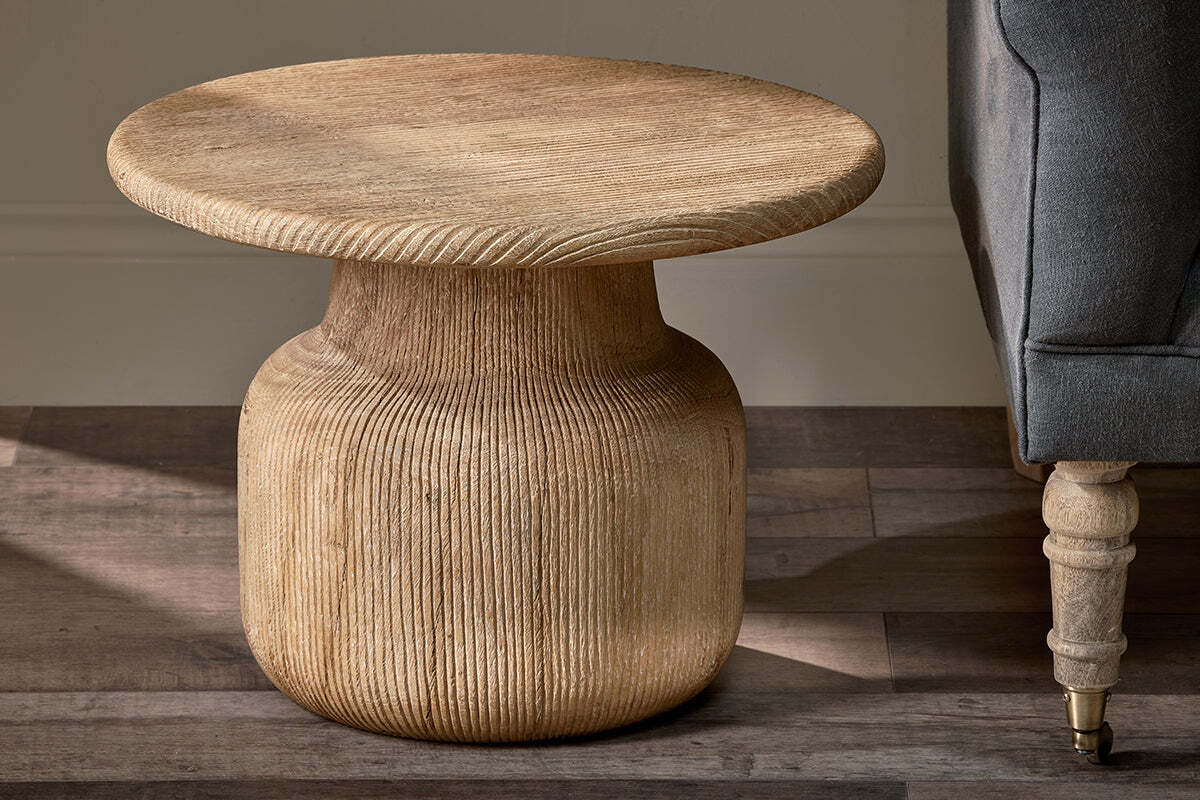 The Vivan Grooved Wood Side Table by nkuku features a round, light mango wood top and textured ribbed base. Sunlight highlights its natural grain as it stands beside a gray upholstered sofa on a wooden floor.