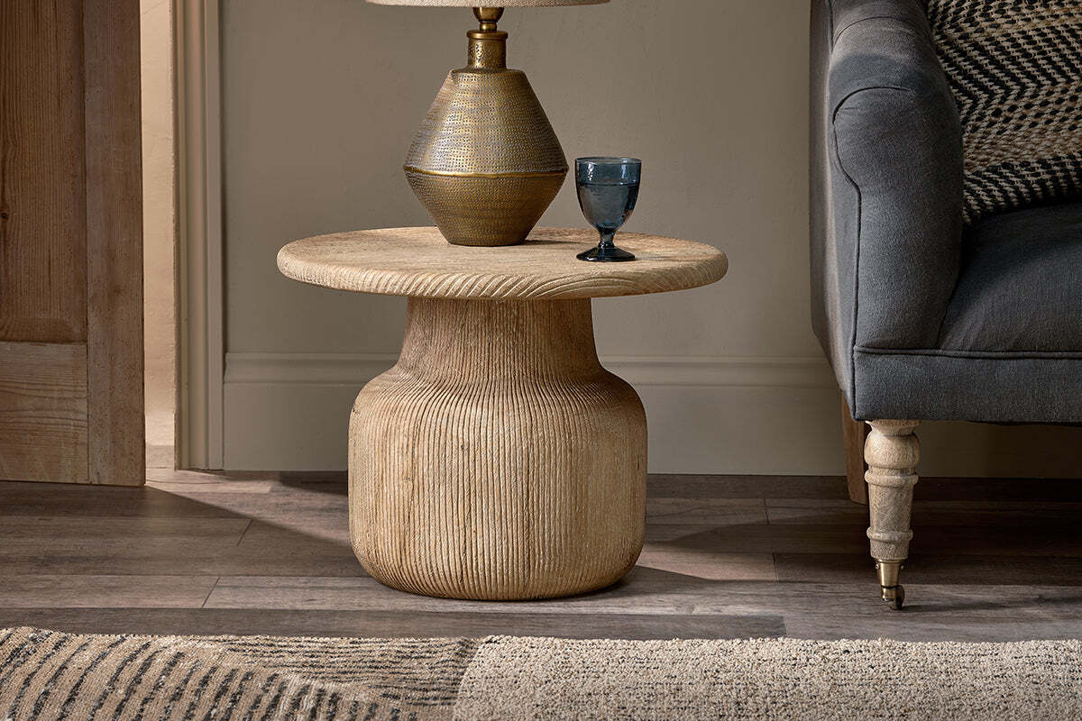 A round, textured Vivan Grooved Wood Side Table by nkuku, made from sustainable mango wood, supports a gold lamp and blue glass beside a gray upholstered armchair on wood flooring, against a light beige wall and door.