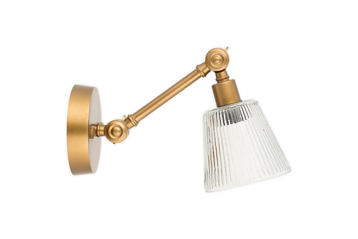 The nkuku Davika Ribbed Glass Task Wall Lamp in clear and antique brass features an adjustable arm and a ribbed glass cone-shaped shade, shown isolated on a white background.