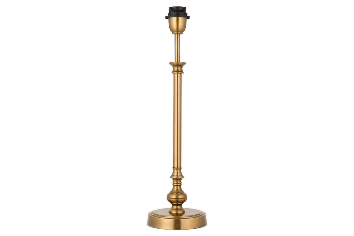 The Ehadi Antique Table Lamp by nkuku features a tall, slender antique brass base with decorative detailing and a round pedestal, shown without shade or bulb, set against a white background.