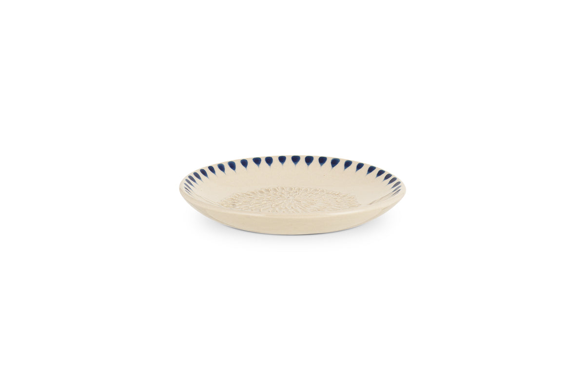 The nkuku Indigo Drop Garlic Grater in Cream & Indigo is a shallow, round beige ceramic plate with a textured center and hand-painted dark blue triangle accents around the rim, perfect for grating garlic.