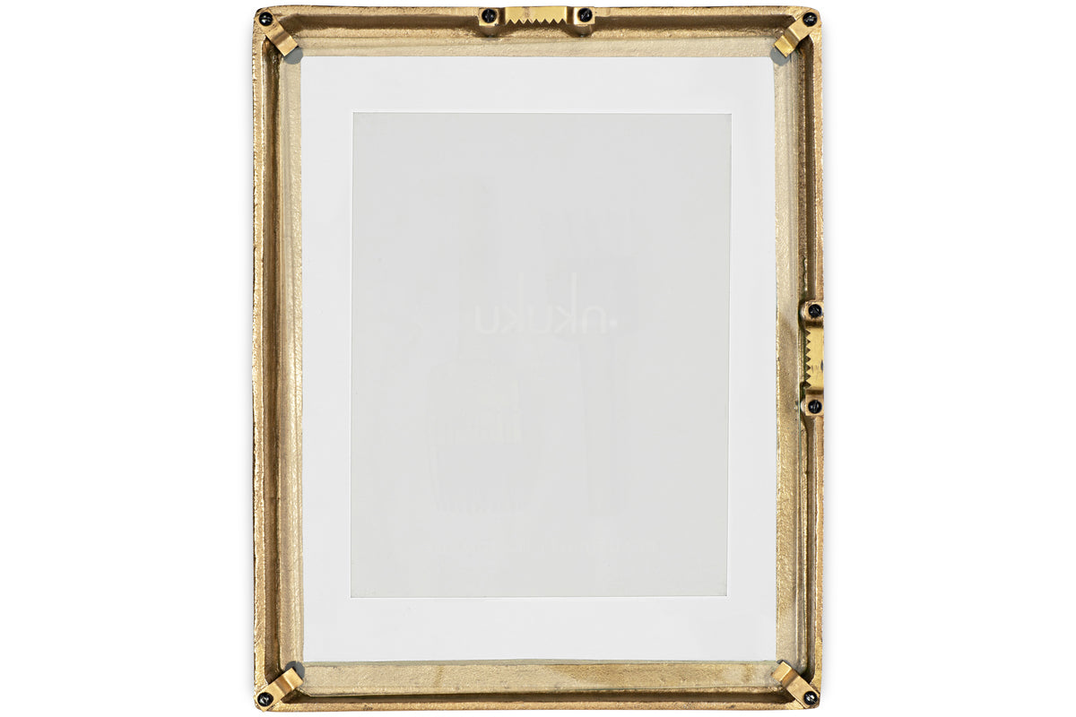 The Jahala Wall Hung Photo Frame by nkuku features a rectangular antique brass frame with decorative ridges and black screws at the corners and sides, and includes a white mat. Shown empty against a white background.