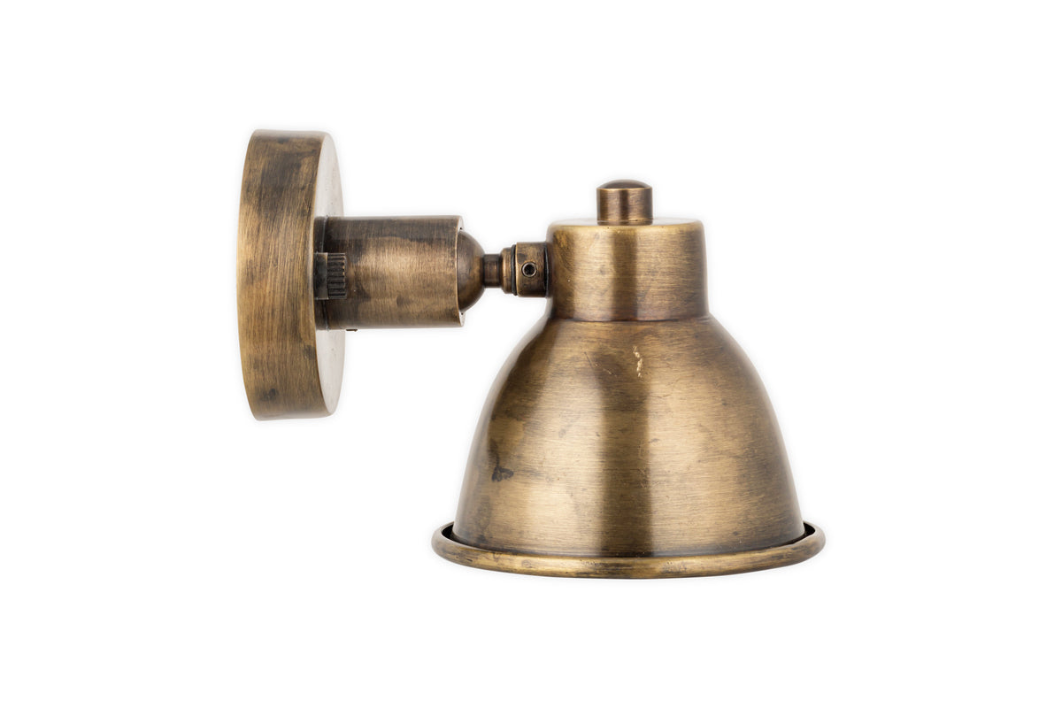 The nkuku Kasai Brass Spotlight in Antique Brass features a rustic, weathered dome-shaped shade, round base, and adjustable arm. This vintage-inspired wall-mounted sconce is shown against a white background.