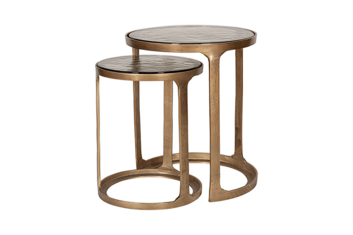 The nkuku Khidki Cast Metal & Glass Nesting Side Tables in antique brass feature two round tables with open bases; the smaller slides under the taller. Both have textured tops and a modern design. Set of 2.