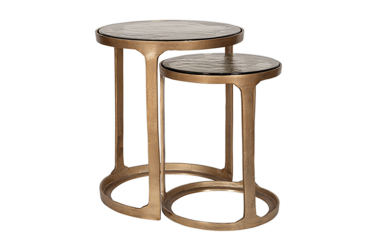 The nkuku Khidki Cast Metal & Glass Nesting Side Tables (Set of 2) feature round glass tops with antique brass-finished metal frames. The smaller table partially nests under the larger, both boasting open circular bases and a modern, minimalist look.