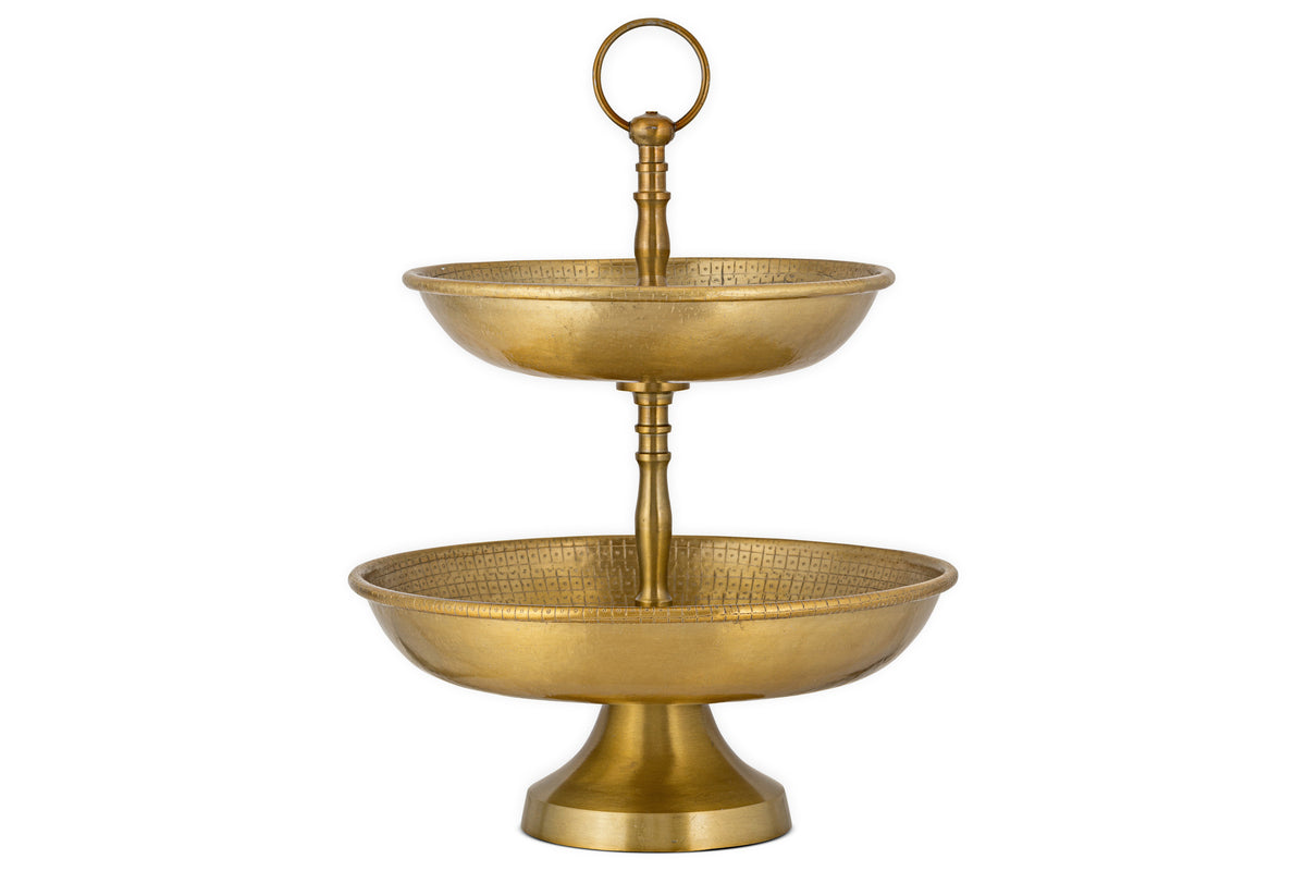 The Mahika Stand - Antique Brass by nkuku is a two-tiered gold metal stand with textured bowls and a top handle, ideal as a centrepiece for festive tablescapes or for displaying fruits, desserts, or snacks.