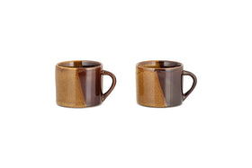 The Naav Mugs - Mustard - Small (Set of 2) by nkuku, each with an angled handle and a glossy mustard and tan color-blocked glaze, are arranged side by side on a white background.
