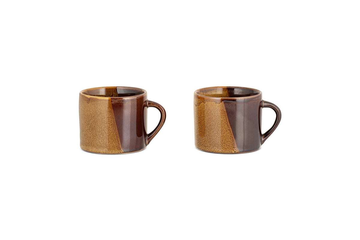 The Naav Mugs - Mustard - Small (Set of 2) by nkuku, each with an angled handle and a glossy mustard and tan color-blocked glaze, are arranged side by side on a white background.