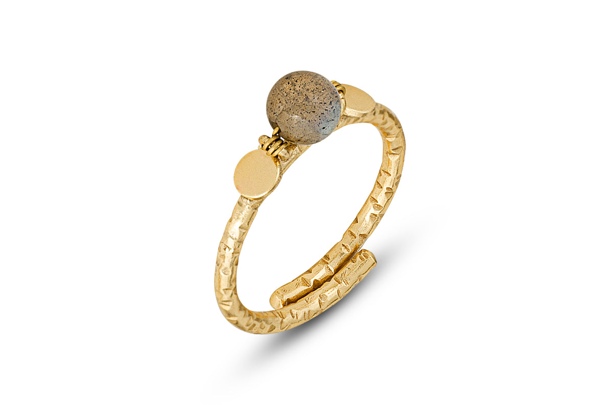 The Marai Labradorite Ring by nkuku features a textured gold-plated adjustable band with a rough, round gray labradorite center, flanked by two flat gold discs. Its hammered finish creates an elegant look, showcased on a white background.