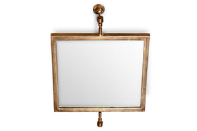 The Visala Swivel Wall Mirror by nkuku features a square, thick antique brass frame and pipe-style supports with exposed hardware, creating an industrial look against a white backdrop.
