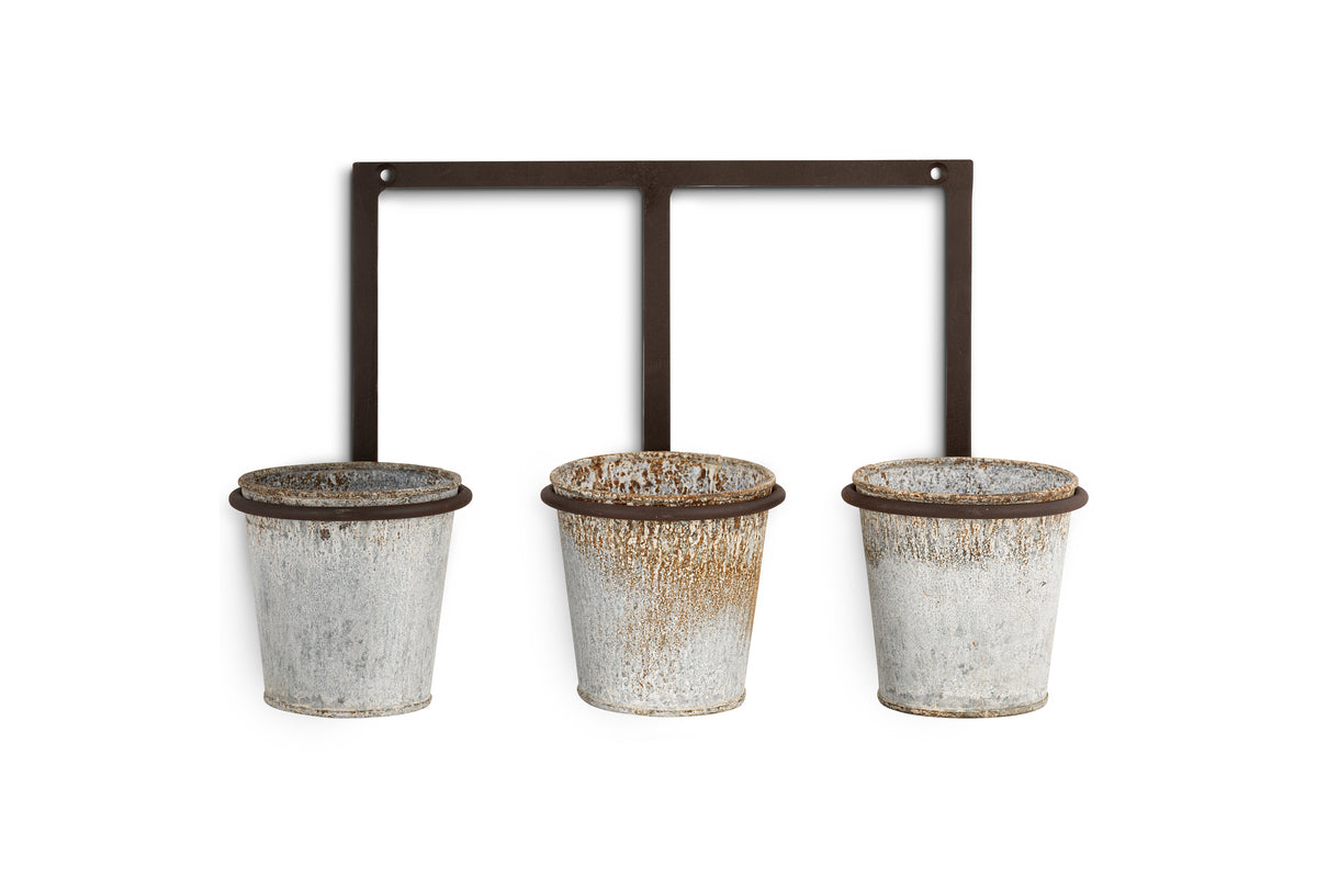 Three small zinc pots with a weathered finish hang from a black metal bar with mounting holes. The nkuku Tukana Triple Wall Hung Planter brings rustic, industrial charm to your space.
