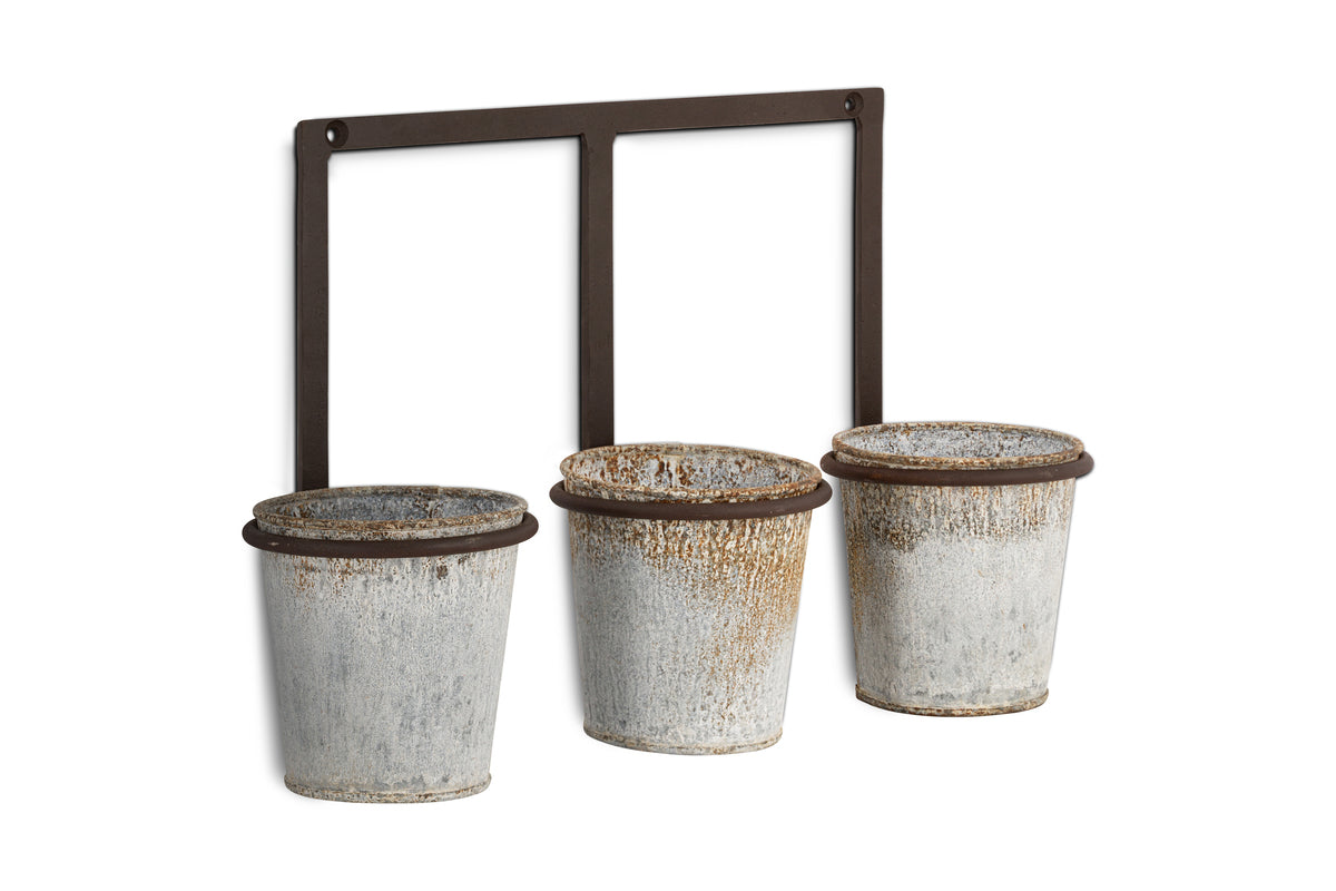 The nkuku Tukana Triple Wall Hung Planter - Zinc features three weathered galvanized pots with rustic patina, each hanging from a ring on a rectangular metal frame for an industrial accent wall display.