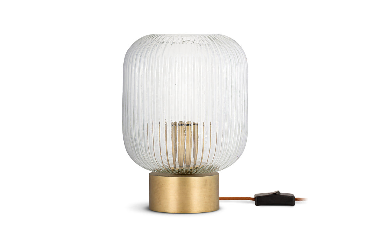 The nkuku Rizal Fluted Glass Bedside Table Lamp in antique brass and clear features a cylindrical ribbed shade, brown cord with black inline switch, and sits atop an elegant brass base against a plain white background.