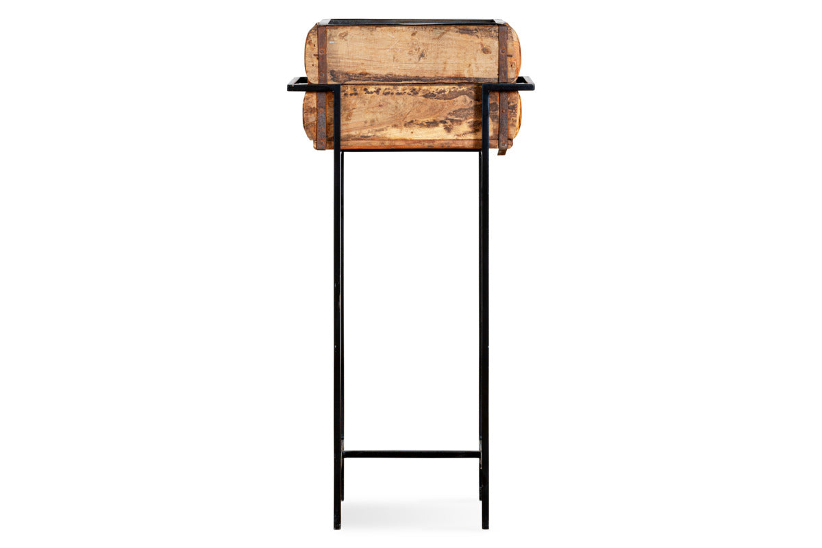 The nkuku Amazzi Indoor Planter Stand - Natural & Black features thin black metal legs and a crossbar, supporting a rustic reclaimed wooden planter box with a weathered finish, shown front-facing against a plain white background.