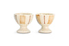 The Patthar Egg Cups - Cream & Clay (Set of 2) by nkuku feature a creamy glaze, vertical tan stripes, and dotted brown lines. Each hand-painted cup has a short, rounded base for added charm. Displayed side by side on a white background.