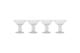 The nkuku Sooraj Champagne Coupe set includes four clear glasses with vintage charm, featuring wide, shallow bowls, short stems, and sturdy round bases. They’re arranged in a row against a plain white background.