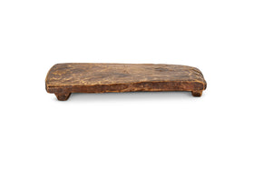 The nkuku Kaveeta Reclaimed Serving Board - Natural is a rustic, rectangular wooden board with rough edges and weathered texture, handcrafted from reclaimed materials. It features four blocky legs and showcases unique wood grain imperfections.